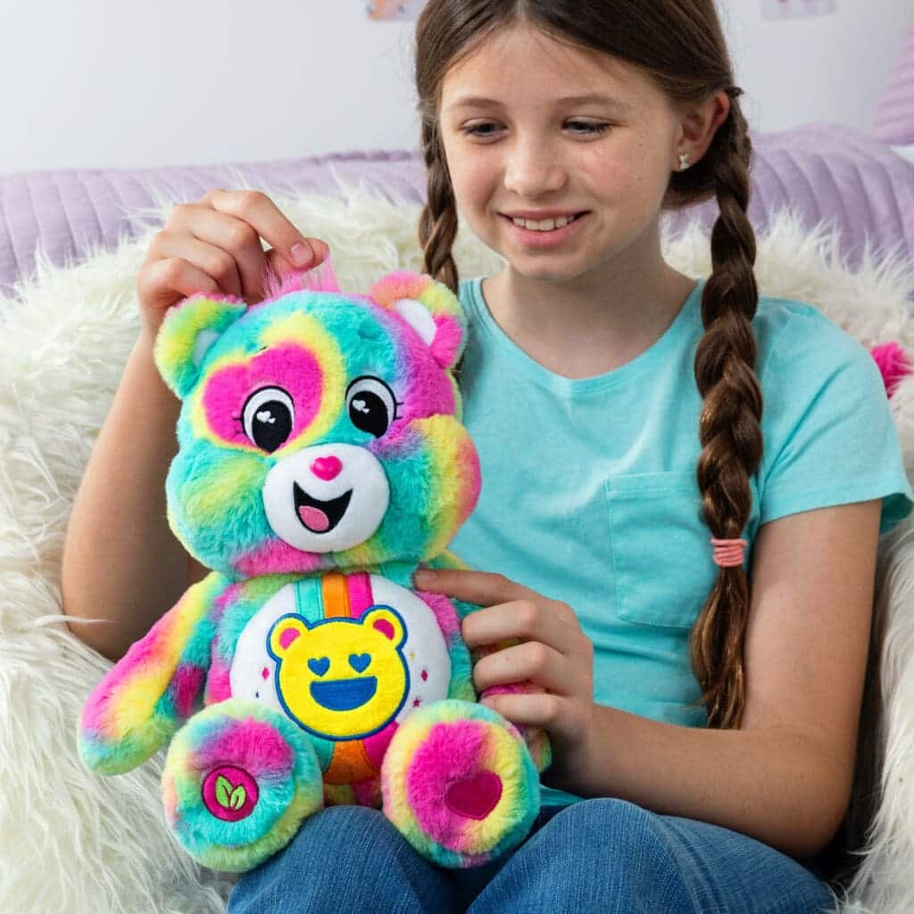 A colorful Good Vibes Bear plush toy from the Care Bears collection is being played with by a young girl.