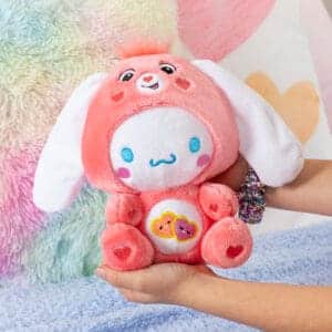 Hands holding Care Bears Funsize Cinnamoroll plush displayed in a lifestyle setting, featuring a cute and cuddly design with a pink heart emblem on its stomach, promoting love and friendship.