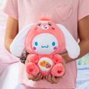 Hands holding Care Bears Funsize Cinnamoroll plush displayed in a lifestyle setting, featuring a cute and cuddly design with a pink heart emblem on its stomach, promoting love and friendship.